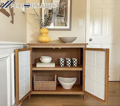 Wooden cabinet with open doors displaying ceramic bowls and a basket, topped with a yellow vase and framed art.